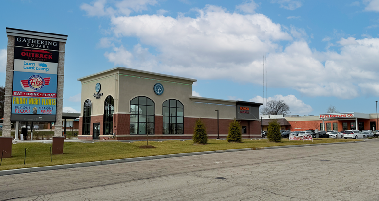 Gathering Square Retail Center entry with the monument sign that shows tenants Outback Steakhouse, Burn Boot Camp and Fitz's. Then a building with Burn Boot Camp and Dunkin Donuts, and ou can see Fitz's Bottling Co. Clouds in a blue sky. 